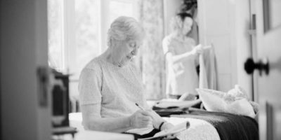 A black and white image of an older lady on a bed writing notes in an assisted living property.